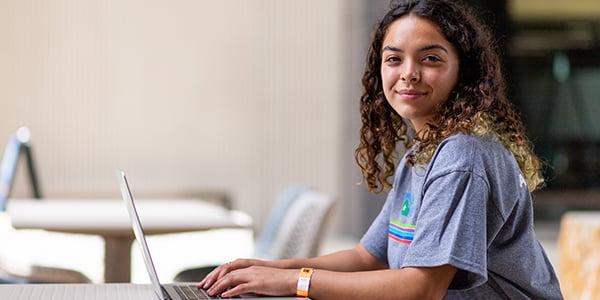 Woman wearing a grey Alamo Promise branded t-shirt while working on a laptop computer